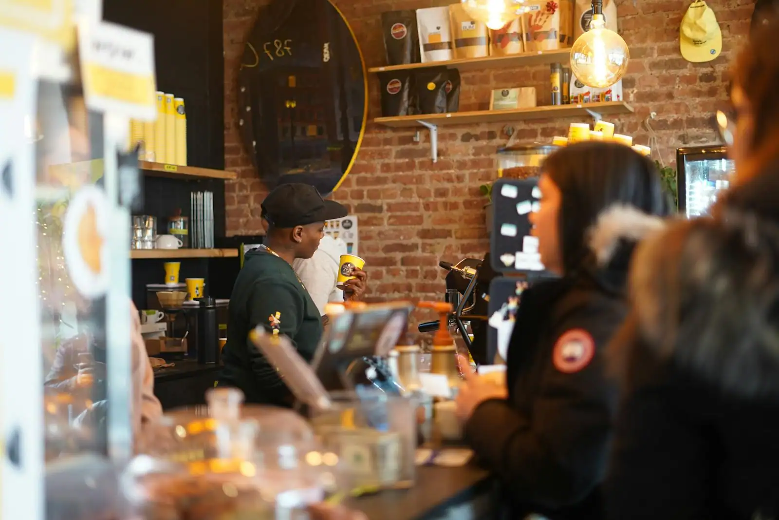 Comptoir d'un café-restaurant avec barista en uniforme servant un cappuccino et clients en arrière-plan, ambiance chaleureuse