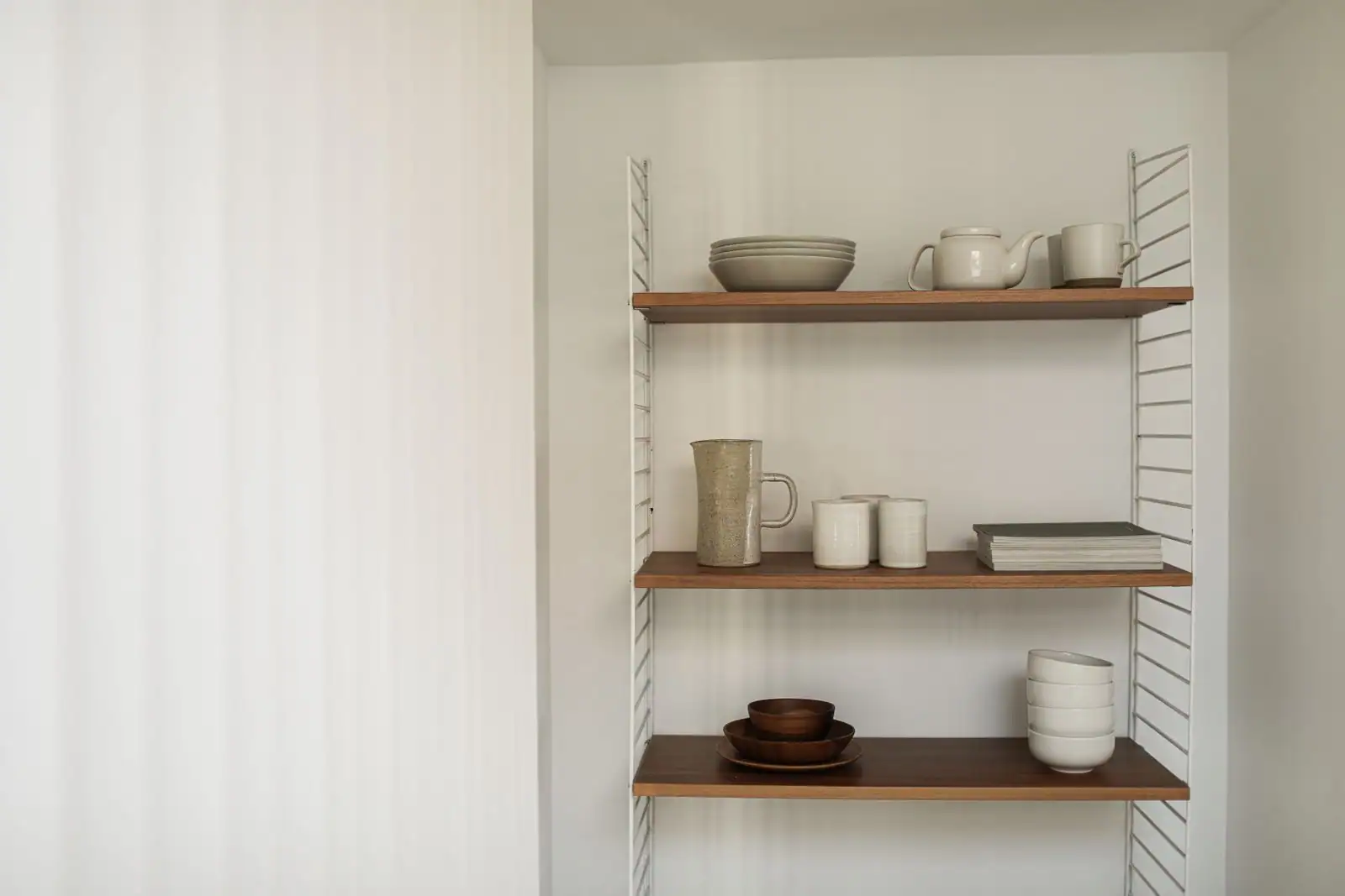 Vaisselle en céramique aux tons neutres — assiettes, bols, tasses et théière — disposée sur des étagères en bois.