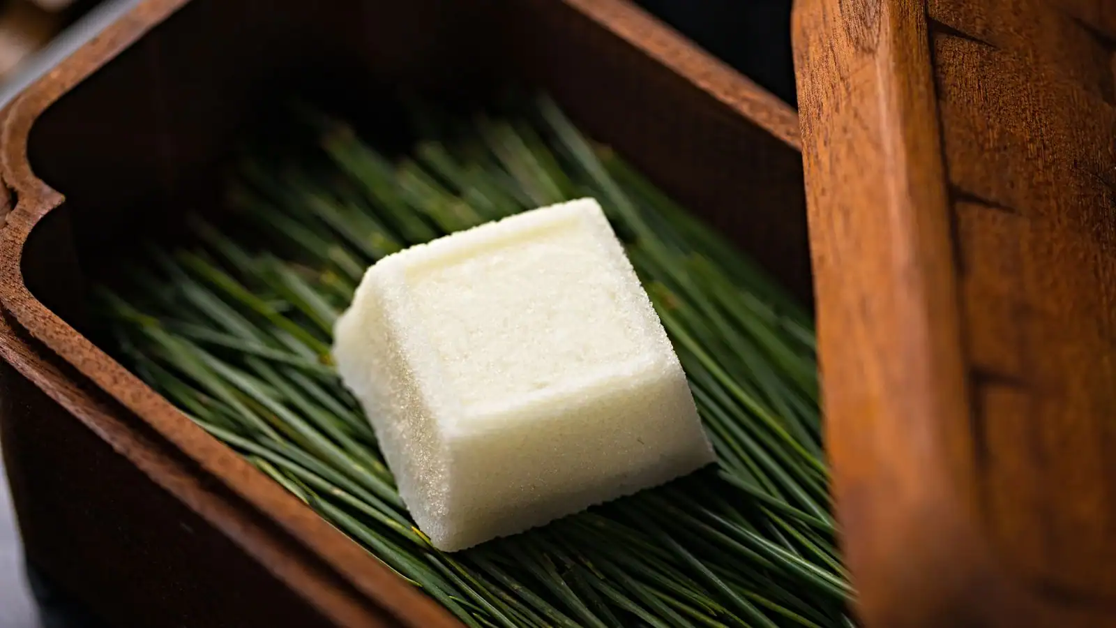 Un wagashi japonais de forme cubique, douceur traditionnelle posée sur des aiguilles de pin dans une boîte en bois.