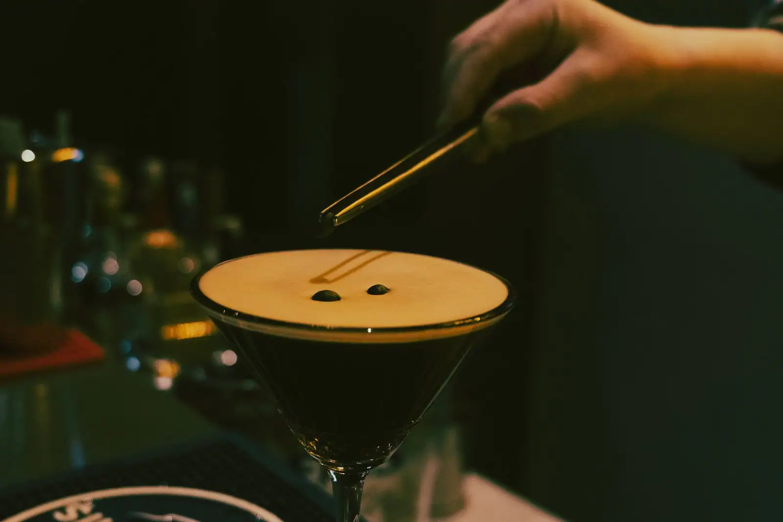 Cocktail Espresso Martini servi dans un verre à pied, coiffé d'une mousse claire, un barman y dépose des grains de café