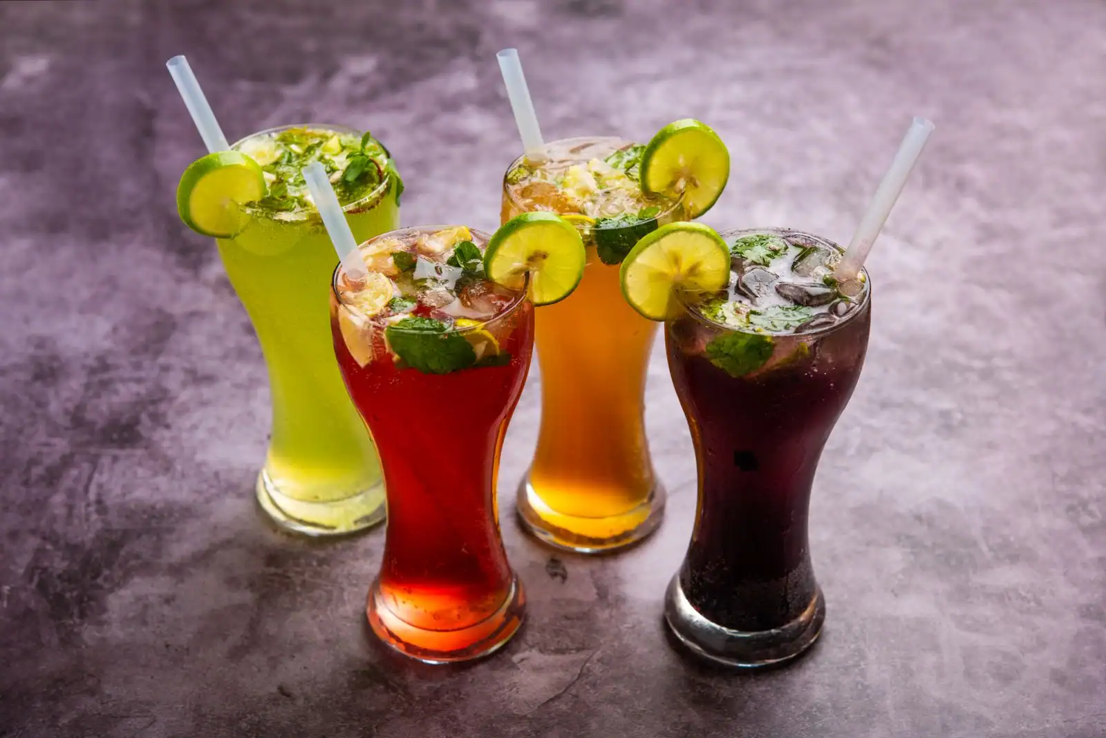 Quatre cocktails sans alcool colorés servis sur glace, garnis de menthe et de rondelles de citron vert.