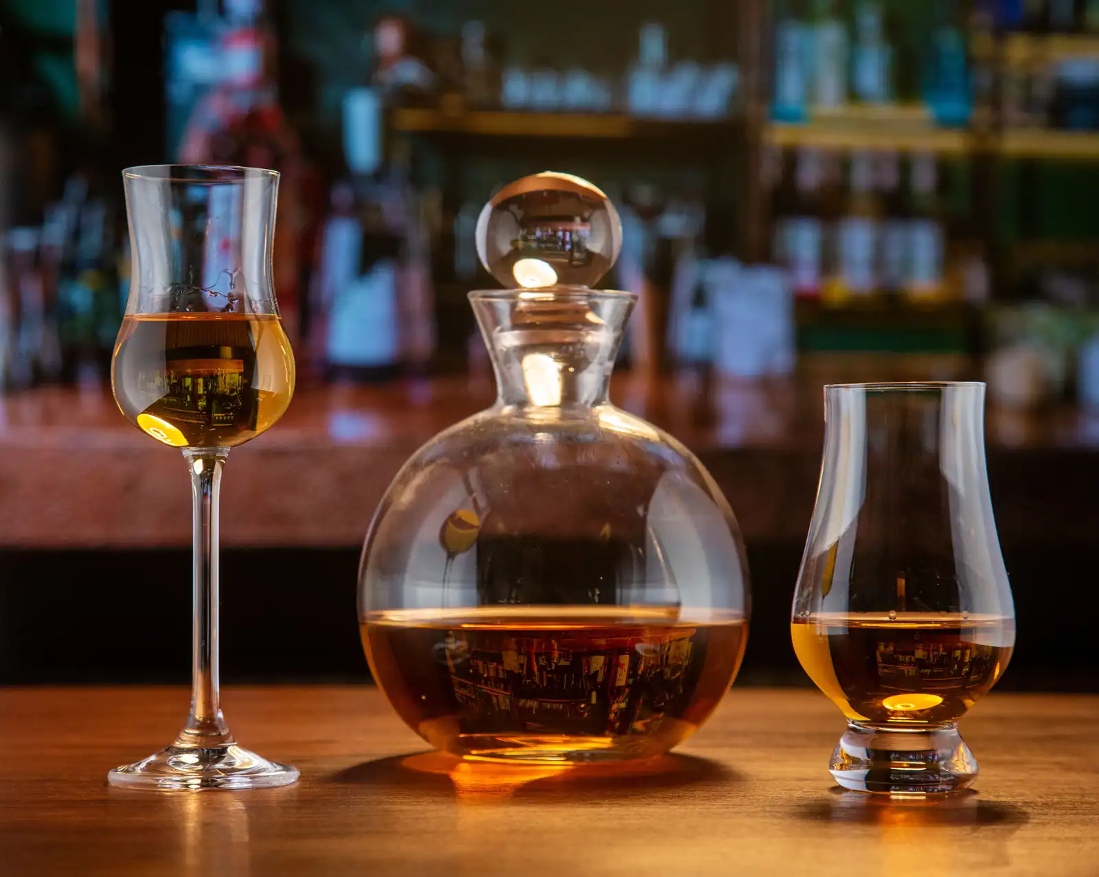 Deux verres tulipe de spiritueux ambré et une carafe, sur un comptoir en bois, dans une lumière feutrée.