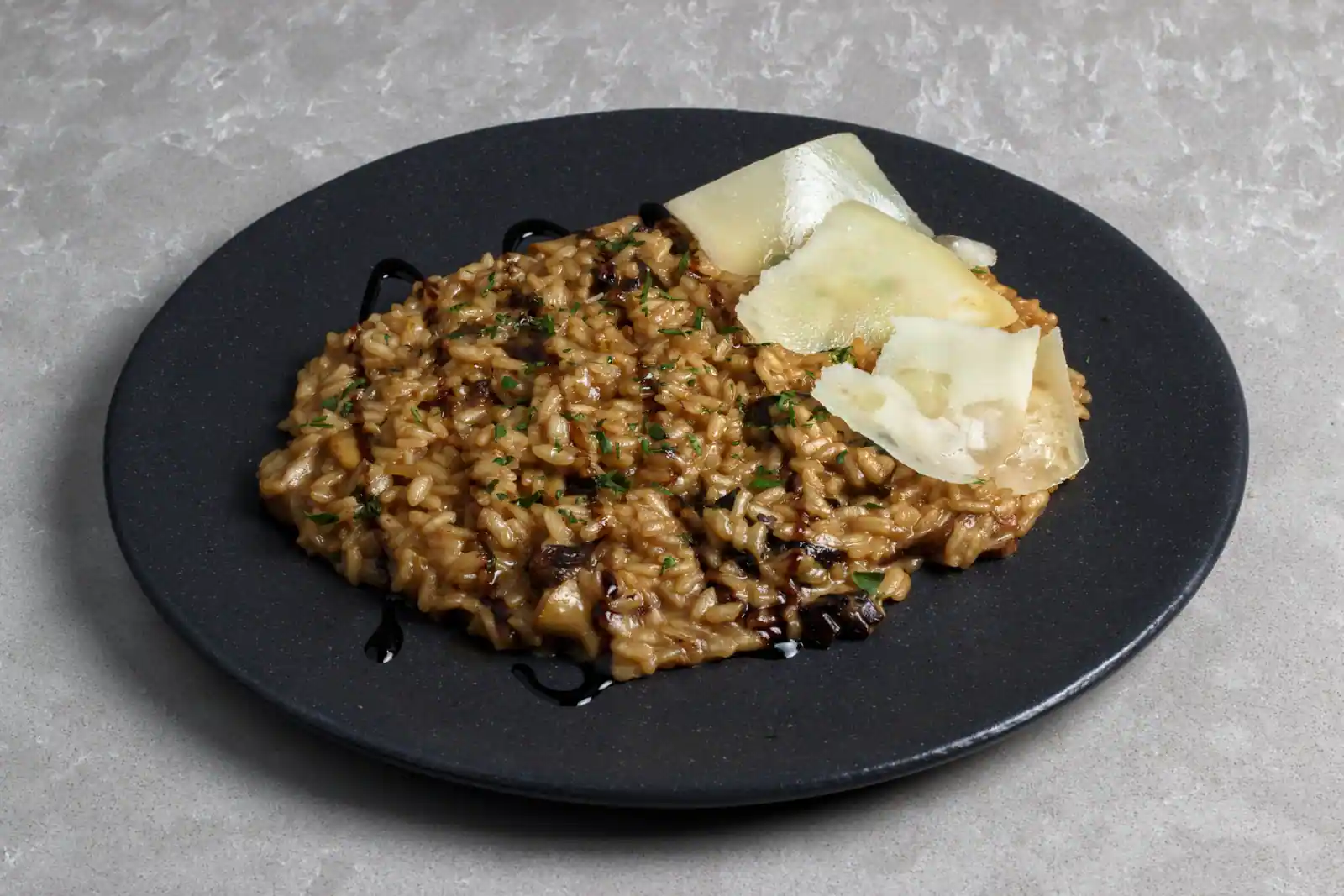 Risotto crémeux aux champignons sur une assiette sombre, garni de copeaux de parmesan et de persil