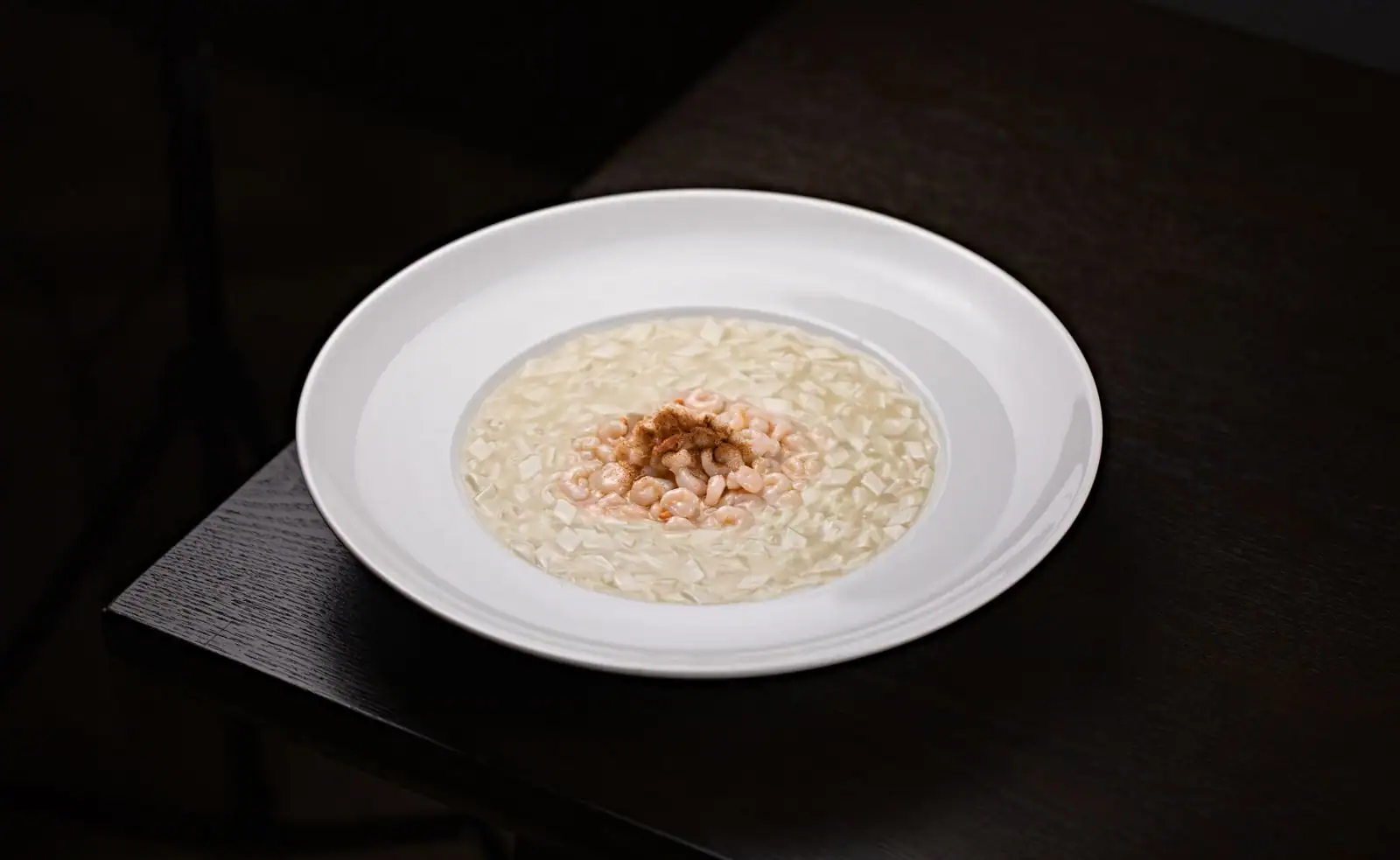 Assiette de risotto crémeux étalé all'onda, surmonté de petites crevettes roses, sur une table sombre.