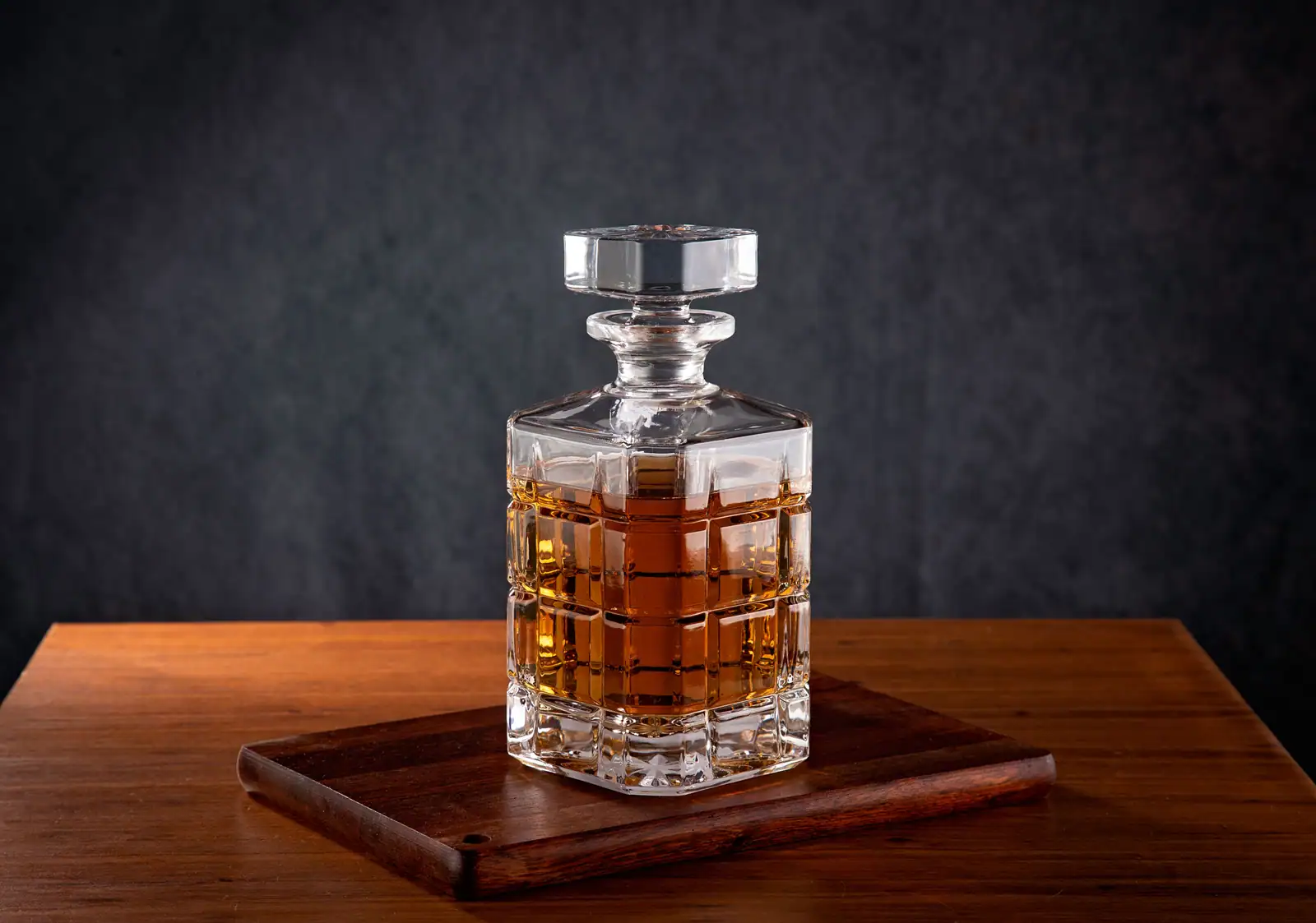 Carafe en cristal taillé remplie de whisky ambré, posée sur une planche en bois sur fond sombre.