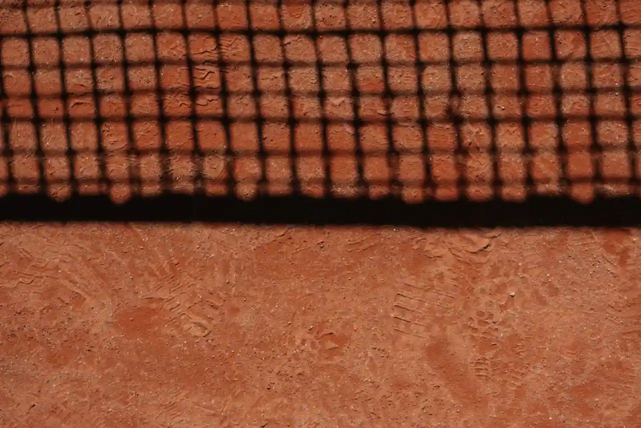 Surface en terre battue rouge d'un court de tennis marquée par l'ombre du filet et des traces de pas