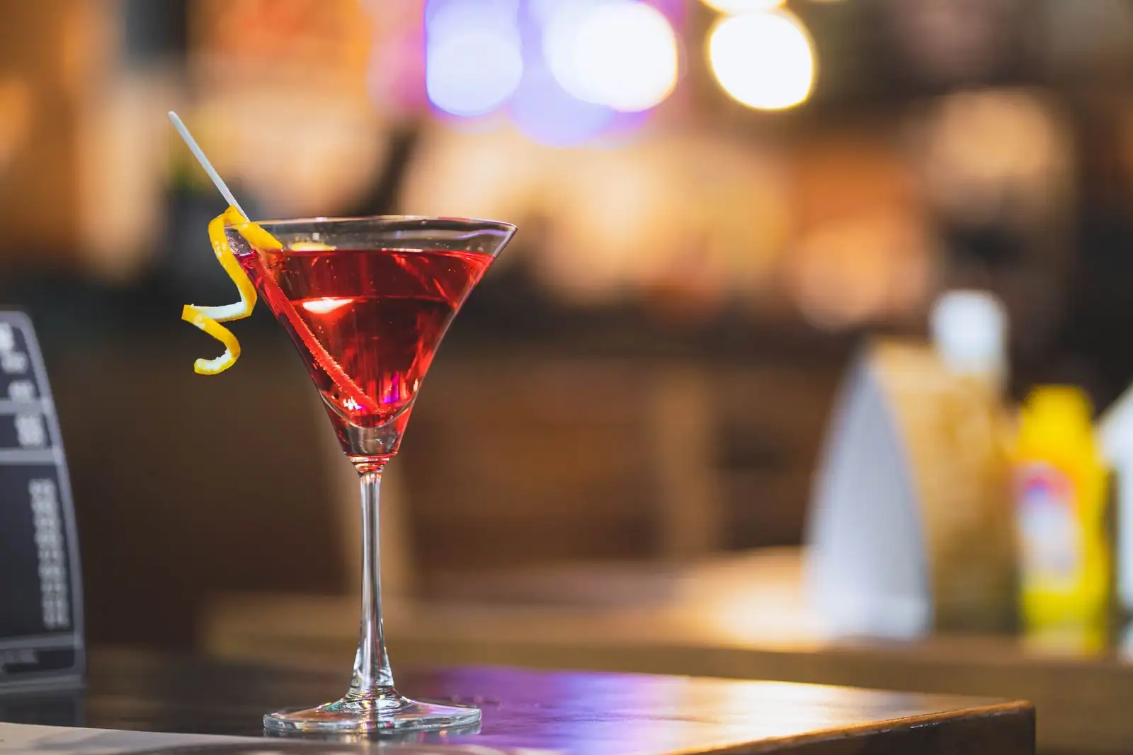 Cocktail rouge et son zeste de citron servi dans un verre à cocktail sur un comptoir de bar, ambiance feutrée