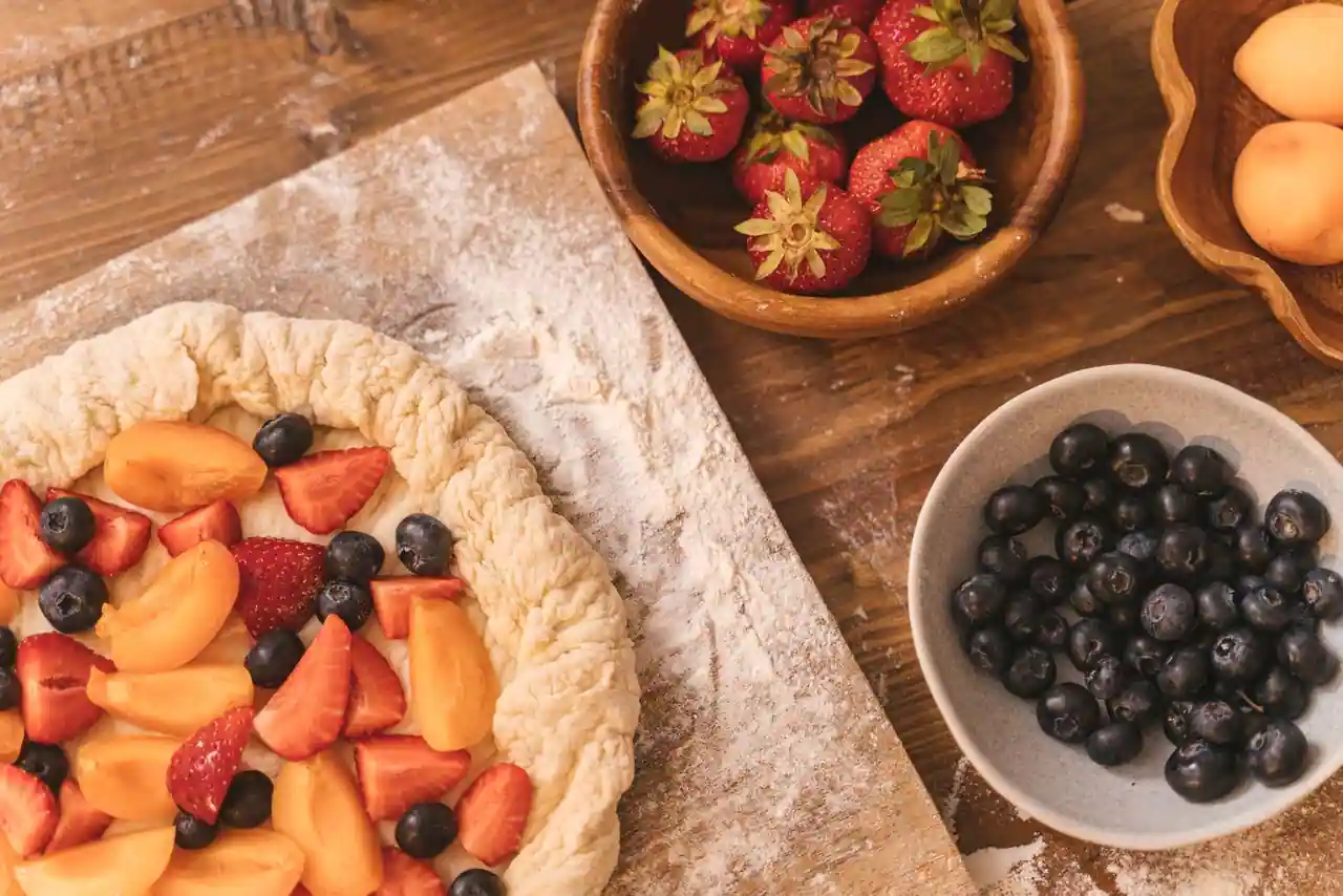 Tarte aux fruits maison en préparation, entourée de fraises, abricots et myrtilles sur une table en bois.