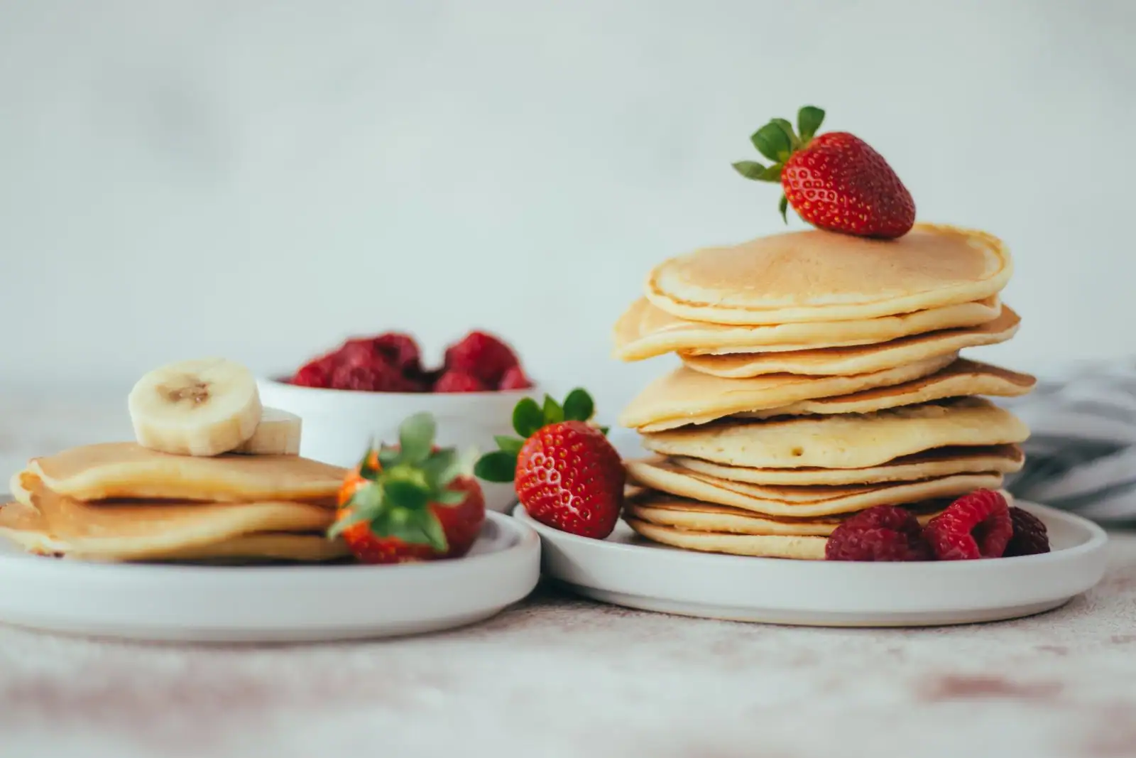 Pile de pancakes dorés surmontée d'une fraise, avec framboises et rondelles de banane autour, dans des assiettes claires