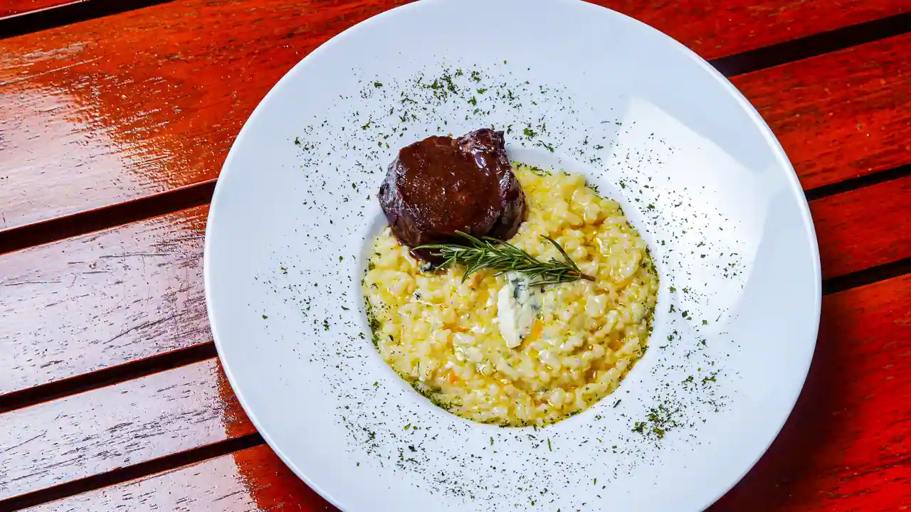Risotto au safran crémeux et doré, servi avec une pièce de bœuf braisé et du romarin dans une assiette blanche