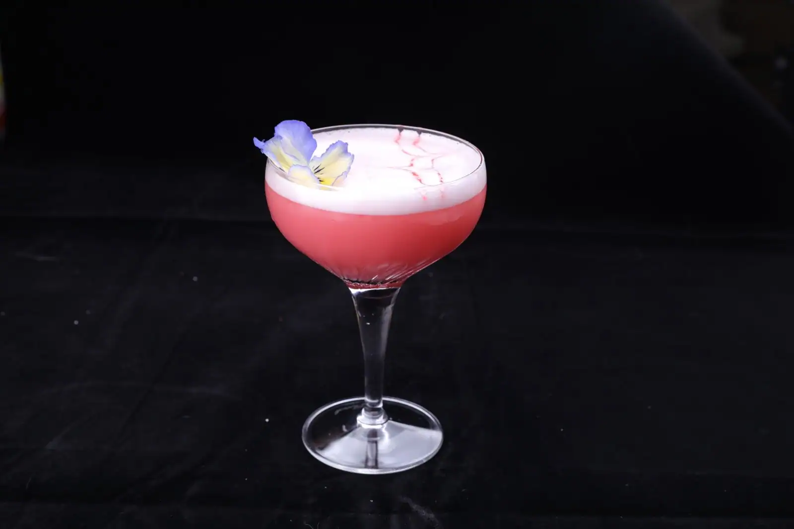 Cocktail rose servi dans un verre coupe transparent, mousse claire en surface et fleur comestible en garniture