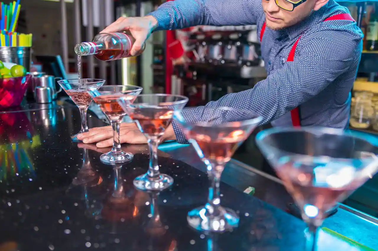 Rangée de verres à cocktail alignés sur un comptoir, remplis un à un derrière le bar pendant le service.