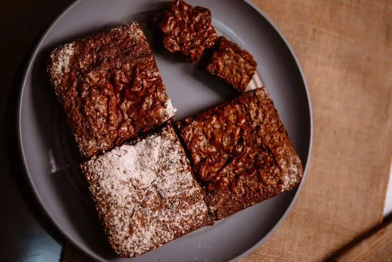 Carrés de brownie au chocolat à la croûte craquelée, saupoudrés de sucre glace, sur une assiette grise.