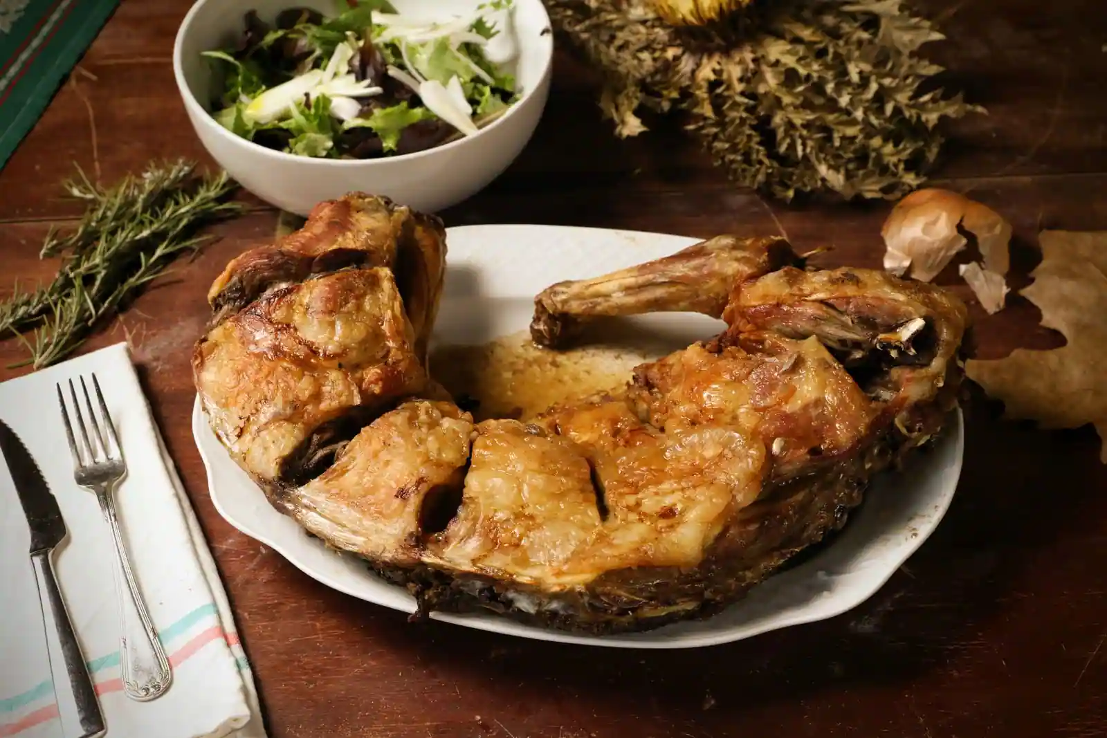 Gigot d'agneau rôti entier servi dans un plat blanc, brin de romarin et couverts sur une table en bois
