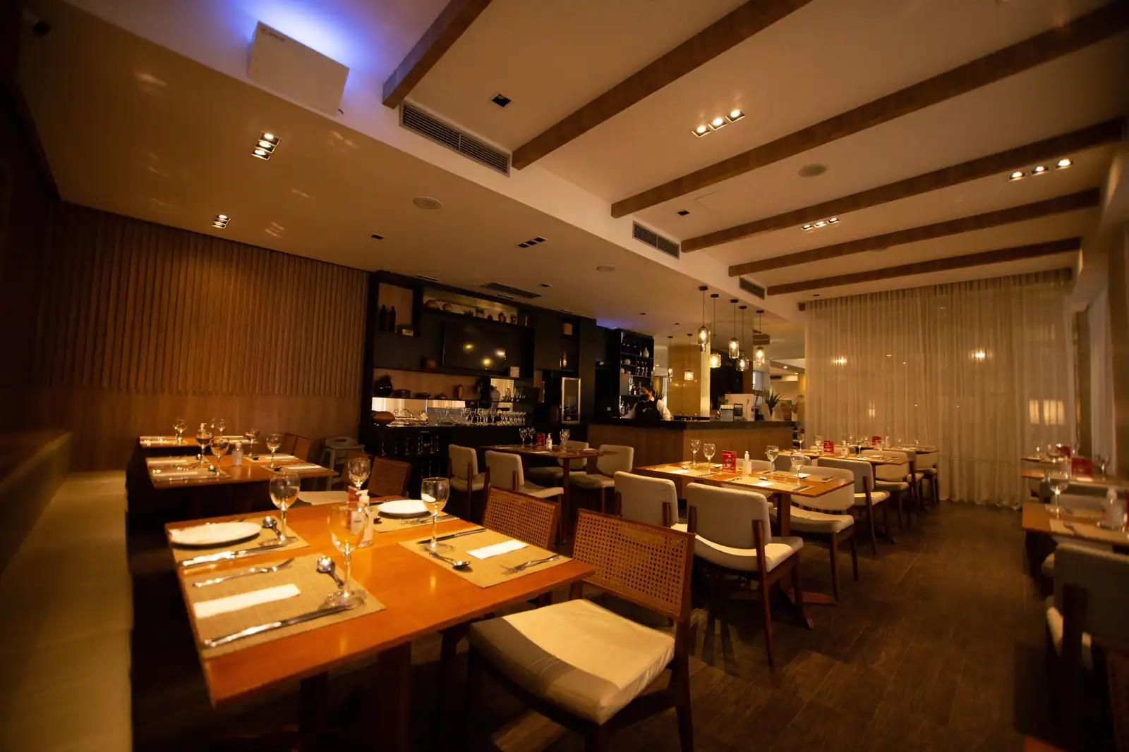 Salle de restaurant vide, tables dressées de verres et de couverts, comptoir éclairé en arrière-plan.