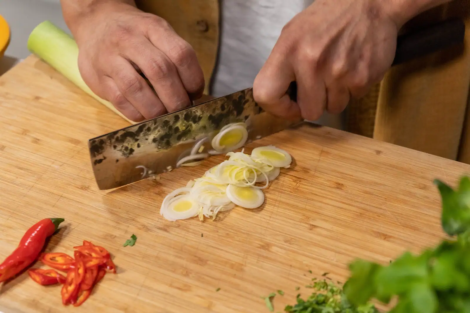 Poireau taillé en rondelles fines au couteau sur une planche en bois, à côté de piment émincé