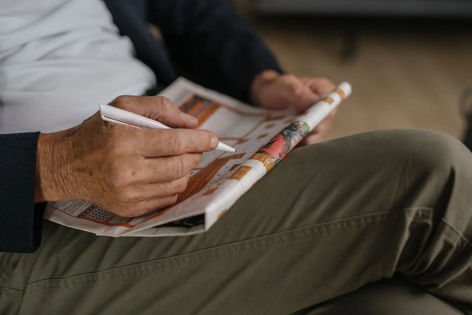 Une main remplit au stylo la grille d'un magazine de jeux de lettres posé sur les genoux
